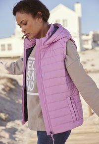 Leichter, bauschiger Weste in Pastellrosa, mit Kapuze, seitlichen Reißverschlusstaschen und horizontaler Steppung. Über einem beigen Langarmshirt getragen.