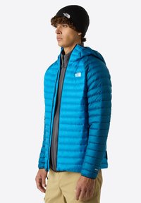 Blå puffjacka med huva, quiltat mönster och grå innerfoder. Svart mössa med logotyp på huvudet. Ljusfärgade byxor.