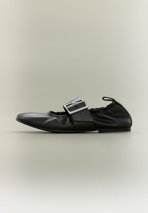 Zapato plano de cuero negro con una gran hebilla plateada, laterales elásticos y una lengüeta trasera, mostrado de perfil sobre un fondo neutro.