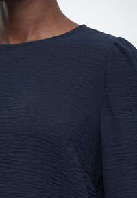 Gros plan d'une personne portant un haut bleu foncé texturé avec un col rond et des manches légèrement bouffantes aux épaules.