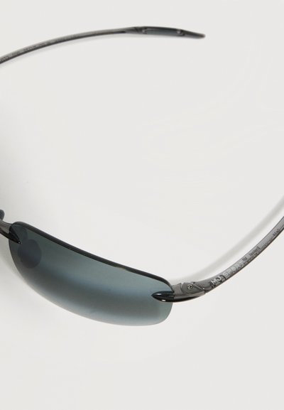 Λεπτά γυαλιά ηλίου χωρίς πλαίσιο με σκούρους φακούς και λεπτούς μεταλλικούς βραχίονες σε απλό φωτεινό φόντο.