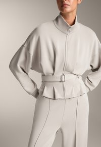 Vaaleanharmaa vetoketjullinen takki, jossa on korkea kaulus, ylisuuri istuvuus, laskostettu helma ja vyötärölle sidottava vyö. Suorapintainen kangas, jossa on vähäiset ompeluyksityiskohdat.