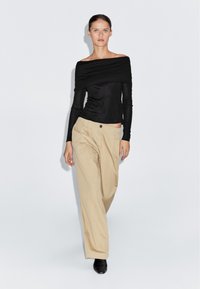 Schwarzes schulterfreies Oberteil mit langen Ärmeln, kombiniert mit beigen, locker sitzenden Hosen und schwarzen High Heels, vor einem einfarbigen Hintergrund.