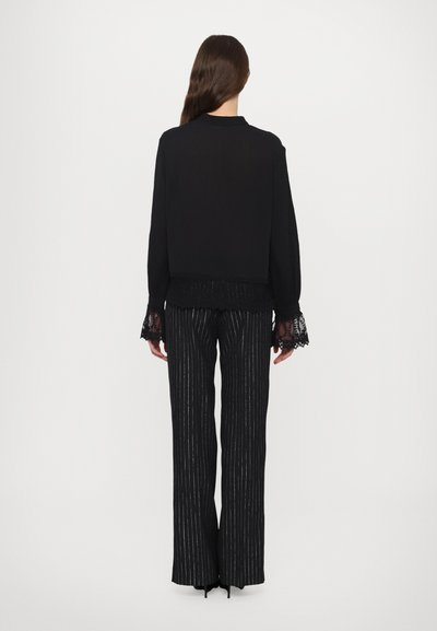Bluză neagră cu mâneci cu dantelă, purtată cu pantaloni largi cu dungi negre și argintii, oferind o textură stratificată și contrastantă.