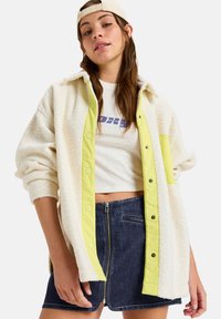 Weiße Fleecejacke mit einer hellgelben Knopfleiste, getragen über einem weißen Cropped-Shirt und einem dunkelblauen Jeansrock mit einem Reißverschluss vorne.