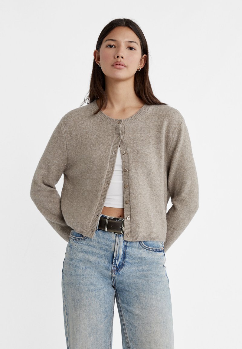 Light brown cardigan made of soft material, featuring a round neckline, front button closure, and long sleeves. Paired with high-waisted jeans.