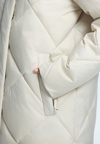 Beige Puffer-Jacke mit gestepptem Muster; verfügt über eine Seitentasche und dicken, strukturierten Stoff für Wärme. Reißverschluss in der Nähe der Tasche sichtbar.
