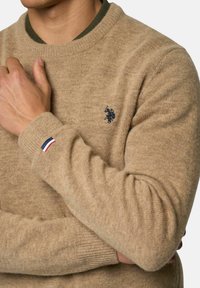 Muž nosiaci béžový pletený sveter s okrúhlym výstrihom a malým námorníckym logom hráča póla na hrudi, s červeným, bielym a modrým detailom na ľavom manžete.