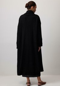 Lungo cappotto di lana nera con davanti aperto, colletto arrotolato e maniche larghe. Presenta un orlo a coste e un tessuto texturizzato, che arriva fino a metà polpaccio.