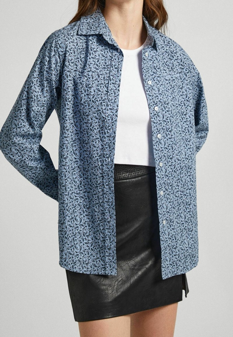 Camisa de manga larga azul claro con un patrón texturizado y cierre de botones. Llevada sobre una camiseta blanca sin mangas y combinada con una falda de cuero negra.