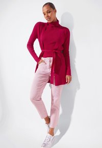 Aansluitende geribbelde coltrui in felrood, gecombineerd met lichtroze gestreepte broek. Afgemaakt met witte sneakers en een gebonden cardigan.