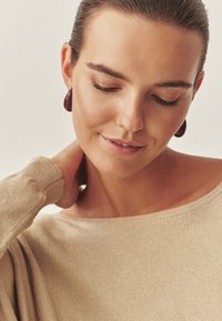 Svetlo bež pleten pulover z izrezom v obliki čolna. Model nosi bordo okrogle uhane, mehko polt in nevtralen make-up. Ozadje z mehkim fokusom.