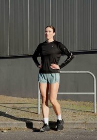 Crna majica dugih rukava za sport, svijetloplave kratke hlače, bijele čarape, crne tenisice. Stojeći uz sivi zid s metalnom ogradom.