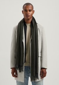 Grijze wollen jas met een notch-kraag, gedragen over een beige trui. Een donkergroene sjaal met franjes hangt losjes om de nek.