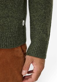 Dunkelgrüner Strickpullover mit gerippten Bündchen und Saum. Verfügt über ein strukturiertes Muster und ein kleines Etikett. Kombiniert mit braunen Cordhosen.