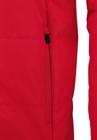 Giacca isolante rossa con zip laterale, texture liscia e design trapuntato. Il materiale sembra leggero e resistente alle intemperie.