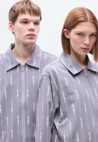 Γκρι ζακέτες με φερμουάρ και τυπωμένο σχέδιο, με κάθετο λευκό κείμενο. Ταιριάζουν σε μοντέρνα σιλουέτα με κλασικό γιακά και άνετη εφαρμογή.