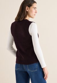 Bordowy, bezrękawnik z teksturowanym splotem i ściągaczem u dołu, noszony na długim białym rękawie, w zestawieniu z niebieskimi dżinsami.