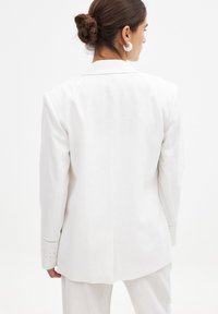 Valge linane bleiser, millel on kohandatud disain, tagaküljel lõhiku ja silmadega detailid varrukatel, lisades tekstuuri ja detaili.