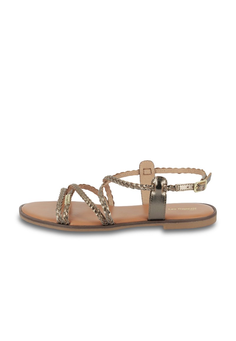 Sandaña plana en dorado metálico con tiras trenzadas. Cuenta con un hebilla ajustable en el tobillo y una plantilla suave en tono marrón claro. Diseño minimalista, estilo abierto en la parte delantera.