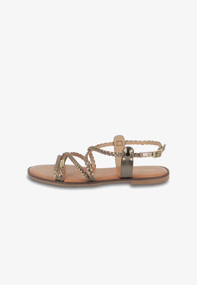 Sandaña plana en dorado metálico con tiras trenzadas. Cuenta con un hebilla ajustable en el tobillo y una plantilla suave en tono marrón claro. Diseño minimalista, estilo abierto en la parte delantera.