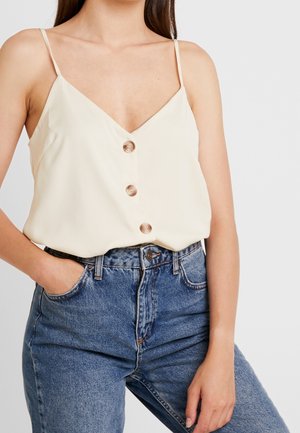 Kobieta w kremowej, bezrękawnikowej bluzce na ramiączkach z trzema guzikami z przodu, włożonej w wysokie niebieskie dżinsy, z ręką w kieszeni.