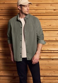 Camisa de botones verde oliva con un bolsillo en el pecho, usada sobre una camiseta beige clara, combinada con pantalones oscuros, con un fondo de madera.