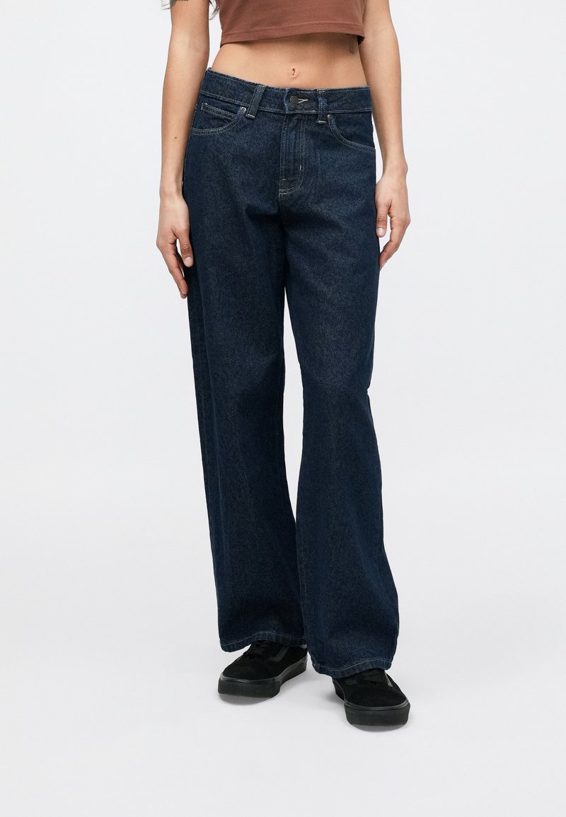 Personne portant un jean denim bleu foncé ample et des baskets noires, debout devant un fond blanc uni.