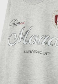 Tissu gris clair avec texte imprimé "Monaco Grand Circuit", un blason brodé rouge et or, et un petit texte "Racing Club" en noir et rouge.