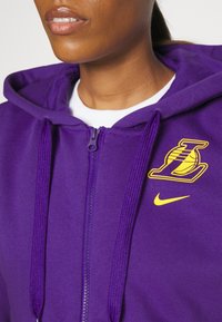 Gros plan d'une personne portant un sweat à capuche violet zippé avec le logo jaune des Lakers de basketball et le swoosh Nike sur la poitrine.