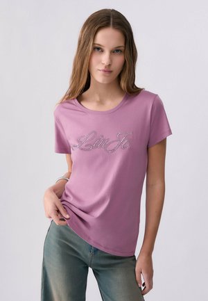 Giovane donna con capelli lunghi indossa una maglietta rosa a maniche corte con "Liu Jo" in strass e jeans blu sbiaditi su uno sfondo semplice.