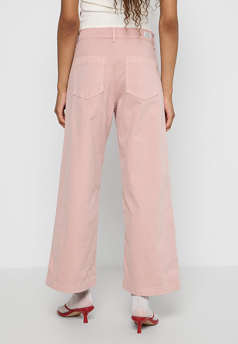 Pantalons à jambes larges en tissu rose doux, avec deux poches arrière et une taille haute. Les finitions des ourlets sont nettes et droites.