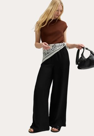 Vrouw met lang blond haar, gekleed in een bruin mouwloos topje, zwarte wijde broekspijpen, witte bandana riem met patroon, en een zwarte handtas vasthoudend.