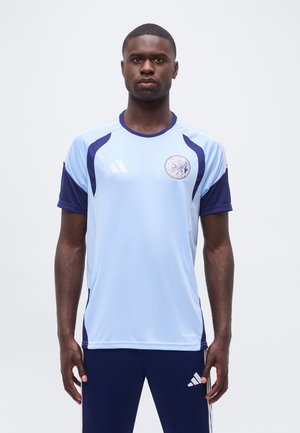 Ein Mann steht nach vorne und trägt ein hellblaues und marineblaues Sporttrikot sowie marineblaue Adidas-Hosen mit sichtbaren Logos auf beiden Kleidungsstücken.