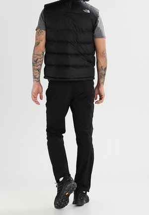 Czarna puchowa kamizelka z wysokim kołnierzem, z przodu na zamek, z logo North Face. Noszona na szarej koszulce, w połączeniu z czarnymi spodniami i butami.