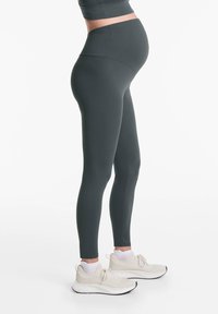 Sötétzöld szövetből készült kismama leggings, magas derékkal, sima textúrával, testhez álló kialakítással. Világos színű tornacipővel párosítva.