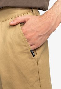 Beige pants with a smooth texture, featuring a side pocket and a black tag labeled "ELEMENT." A hand rests in the pocket.
