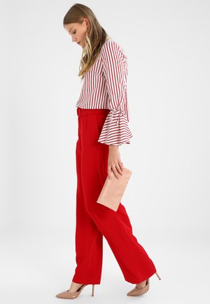 Gestreifte Bluse mit langen Ärmeln und weit ausgestellten Manschetten, kombiniert mit roten, weit geschnittenen Hosen. Das Model hält eine hellrosa Clutch und trägt nude-farbene High Heels.