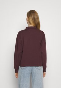 Vínová polokošile s dlouhým rukávem, límečkem, volnějším střihem a žebrovanými manžetami, v kombinaci se světle modrými vysokými džíny.