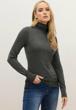 Femme blonde portant un pull à col roulé gris foncé et un jean bleu, une main dans sa poche, sur un fond clair uni.