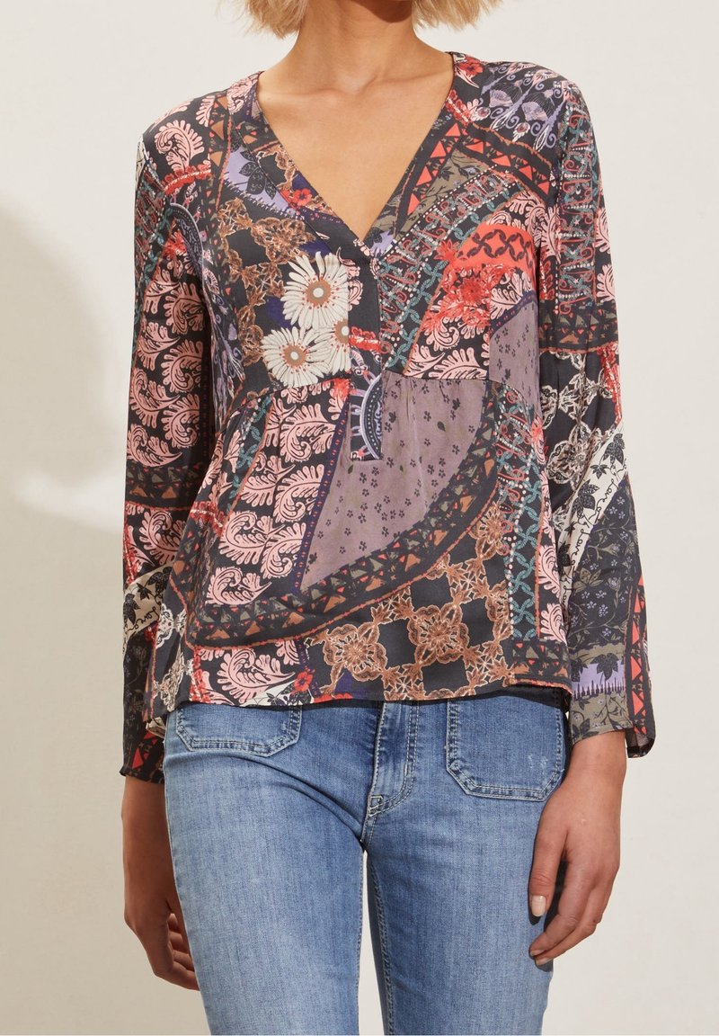 Kvinna iklädd en flerfärgad patchwork-blomblus med långa ärmar och V-ringning, tillsammans med blå denimjeans.