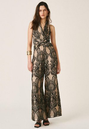 Mujer con mono sin mangas beige con patrón floral negro ornamentado, pantalones anchos, pulseras doradas y tacones abiertos negros, de pie en pose neutral.