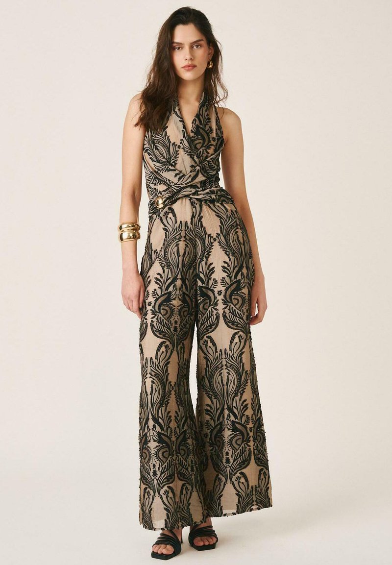 Mujer con mono sin mangas beige con patrón floral negro ornamentado, pantalones anchos, pulseras doradas y tacones abiertos negros, de pie en pose neutral.