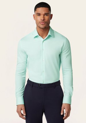 Man draagt een mintgroen overhemd met lange mouwen en knopen, ingestopt in een zwarte broek, staand tegen een effen achtergrond.