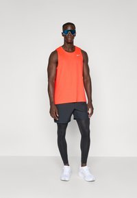 Idrettsmann iført lys orange ermeløs topp, sorte shorts over sorte leggings, hvite joggesko og reflekterende sportssolbriller som står.