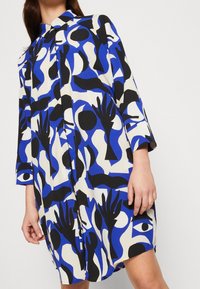 Kraťasové šaty vyrobené z lehké tkaniny, s odvážným geometrickým vzorem v modré, černé a bílé barvě, s límečkem a zapínáním na knoflíky.