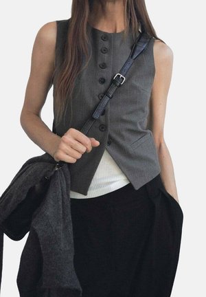 Frau, die eine ärmellose graue, geknöpfte Weste über einem weißen Top trägt, hält einen dunklen Pullover und eine schwarze Umhängetasche, mit einer Hand in der Tasche der schwarzen Hose.