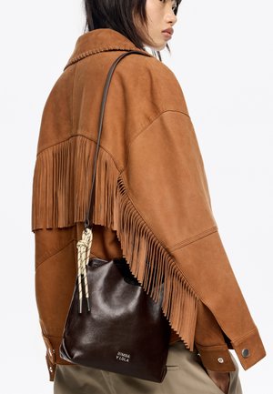Brązowa zamszowa kurtka z frędzlami, noszona na brązowej skórzanej torbie typu bucket z węzłowym paskiem i wytłoczonym logo marki.