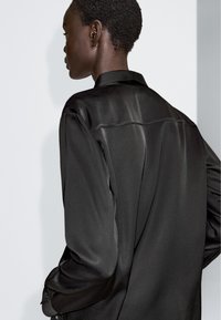 Czarna satynowa koszula o gładkiej fakturze, z długimi rękawami i plisą z tyłu. Posiada klasyczny kołnierzyk oraz mankiety zapinane na guziki.