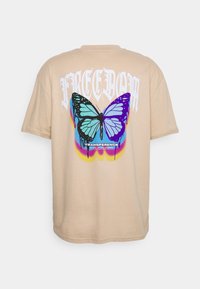 Beige tričko s velkým, barevným grafickým motivem motýla na zadní straně a bílým nápisem "SVOBODA" nad ním. Měkký bavlněný materiál.